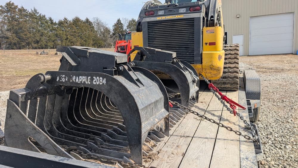 New Holland skid steer with grapple attachment on a trailer at a construction site.