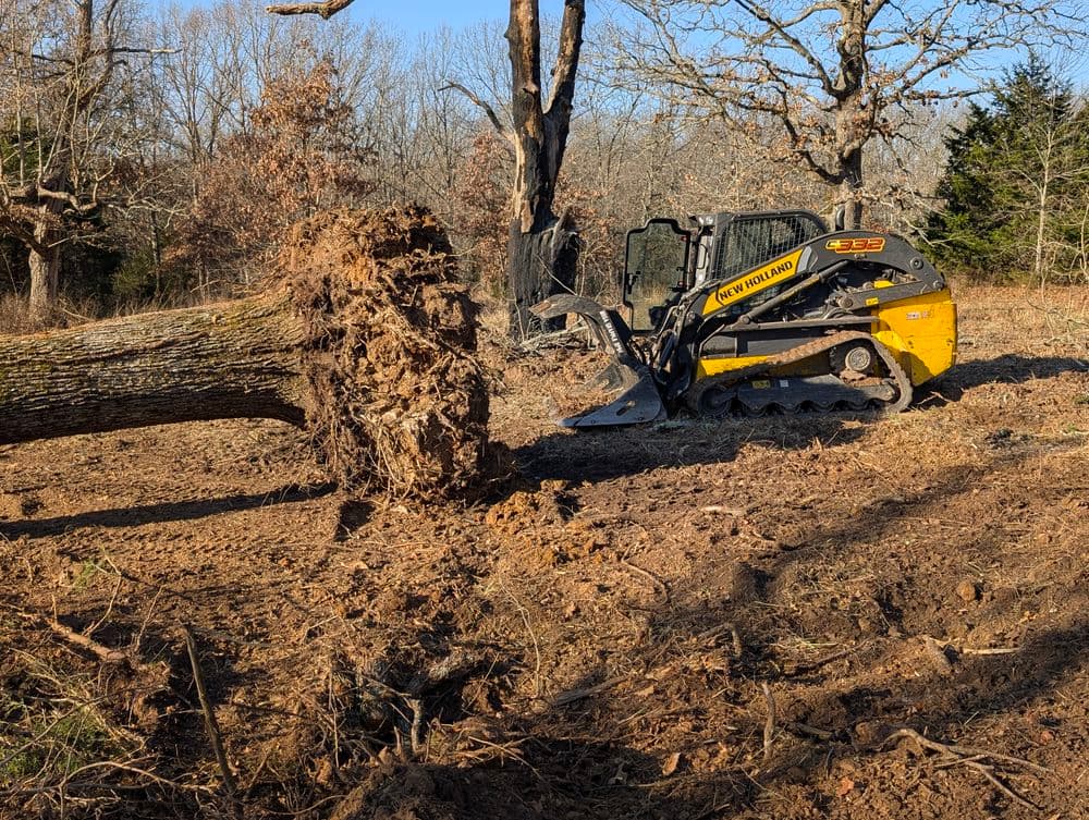 New Holland skid steer loader uprooting a tree in a wooded area.