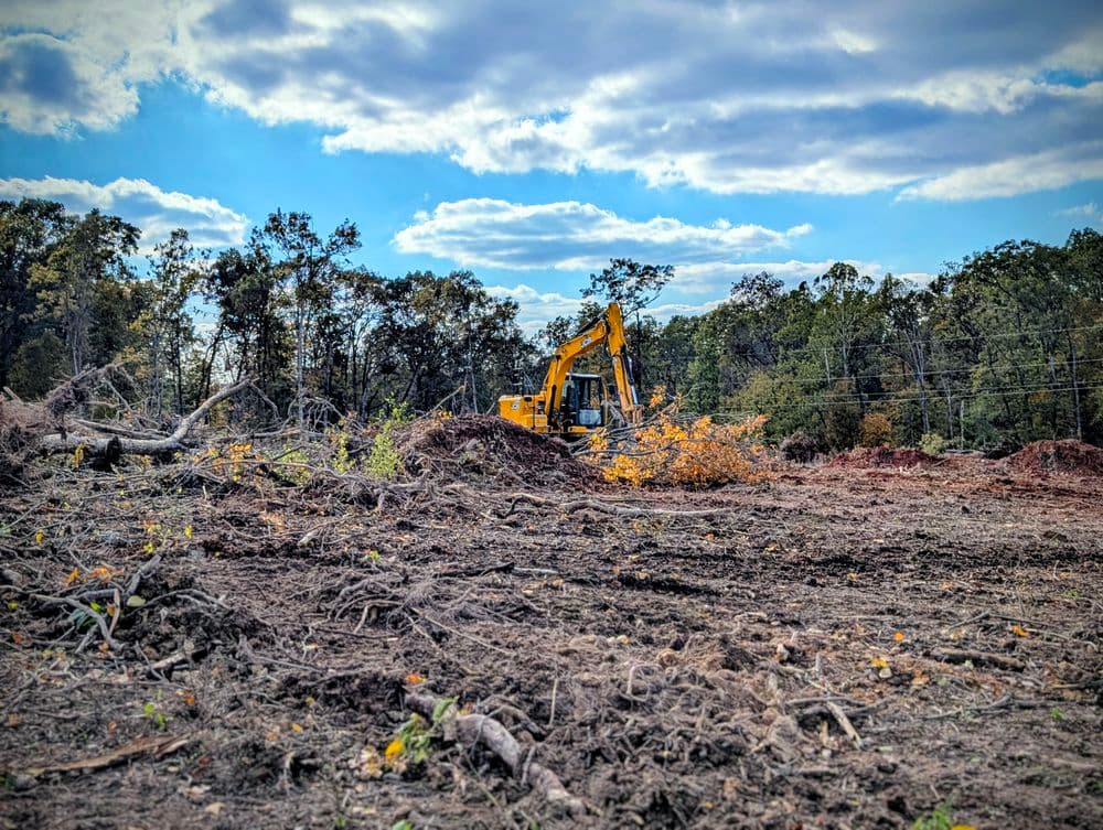 Excavator clearing land with fallen trees under a blue sky and scattered clouds.