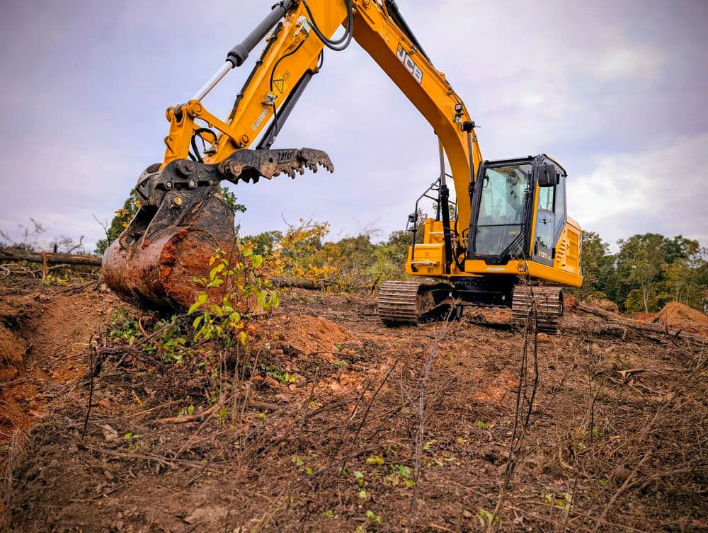 Excavator digging in a cleared land area, surrounded by tree stumps and overcast sky.