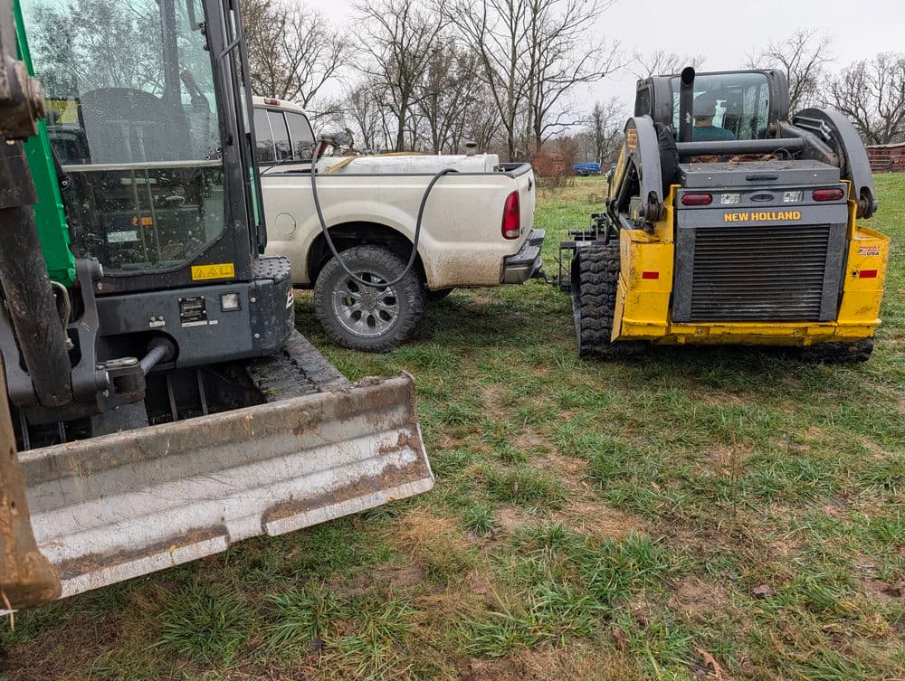 Excavator and skid steer loader next to a pickup truck on a grassy field.