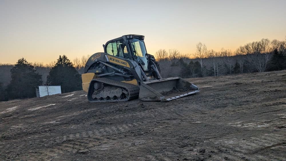 Skid steer loader on a construction site with trees and sunset in the background.