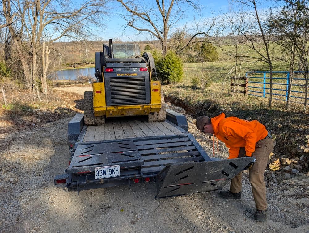 Skid steer loader on trailer with operator adjusting ramp near rural landscape and pond.