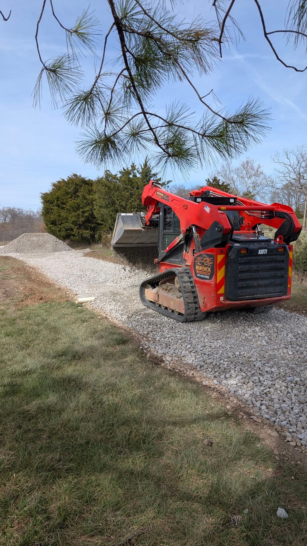 Excavator spreading gravel on a driveway surrounded by trees and grass.