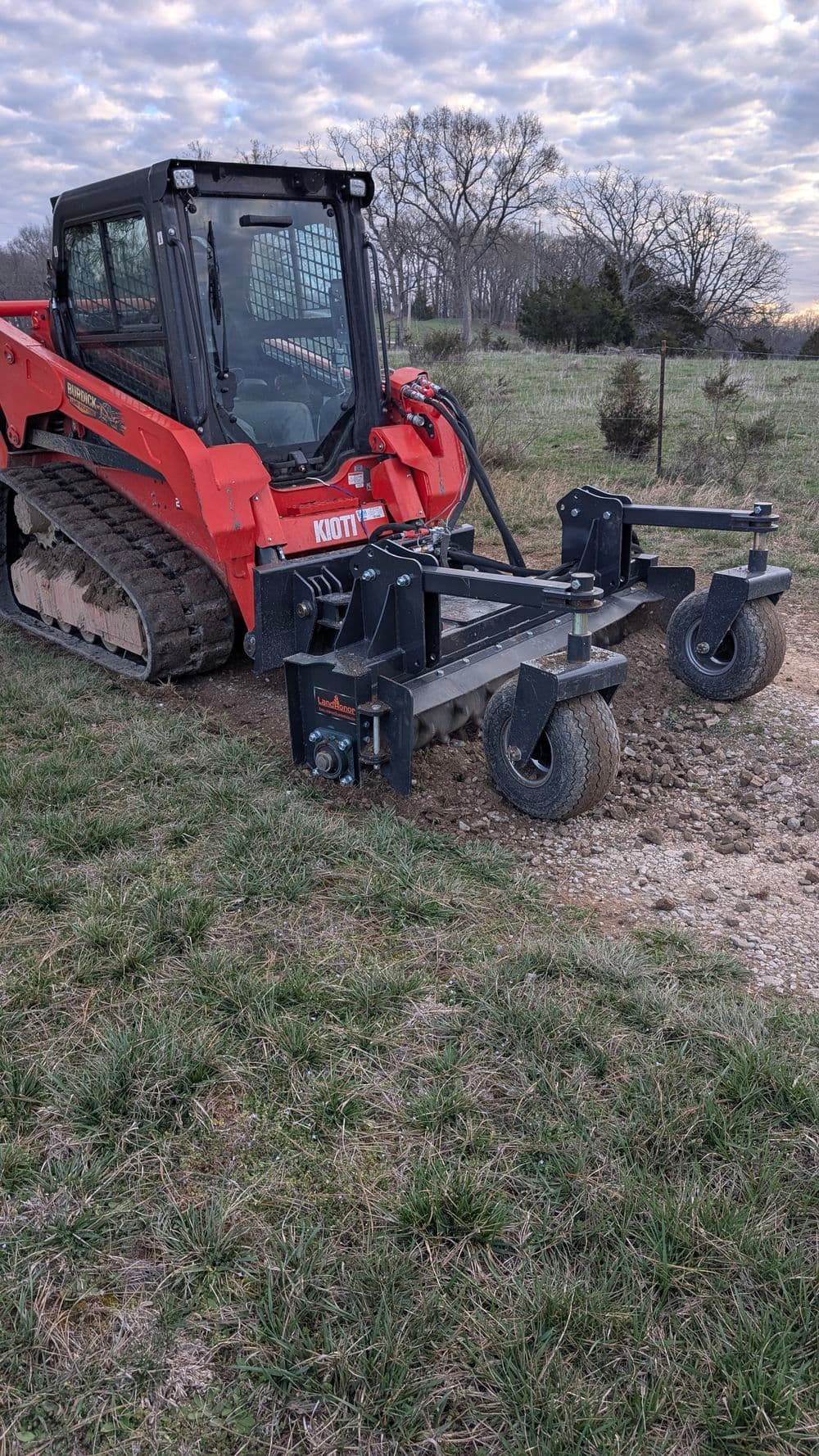 Red skid steer loader on gravel drive, featuring a heavy-duty grading attachment.