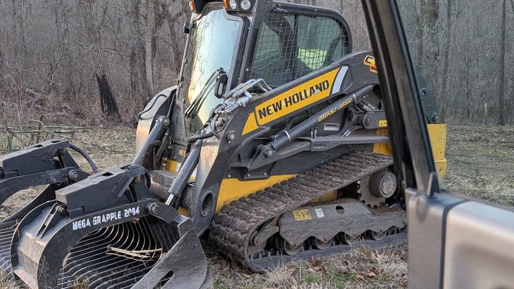 New Holland skid steer loader with a mega grapple attachment in a forest clearing.