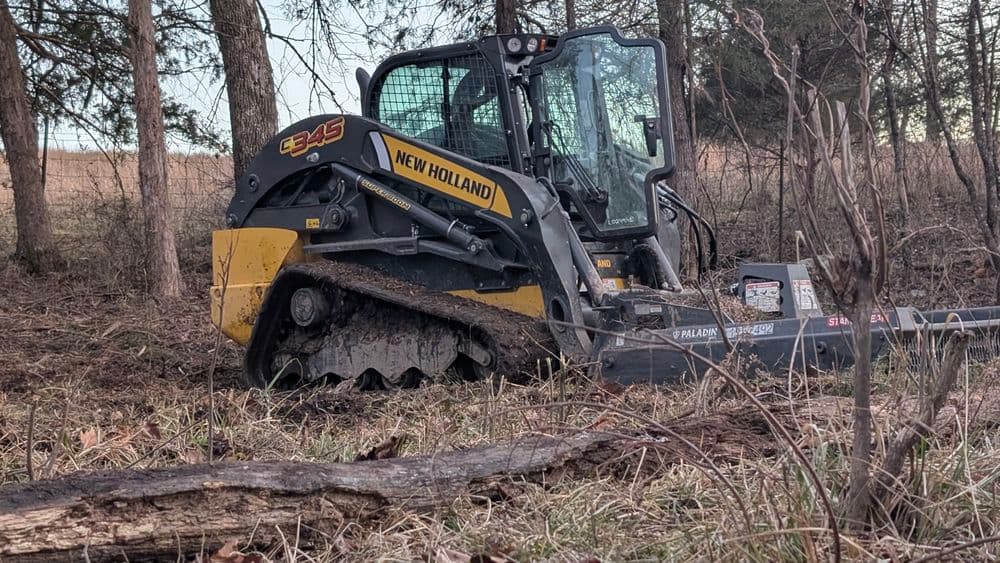New Holland 345 skid steer loader clearing brush in a forested area.