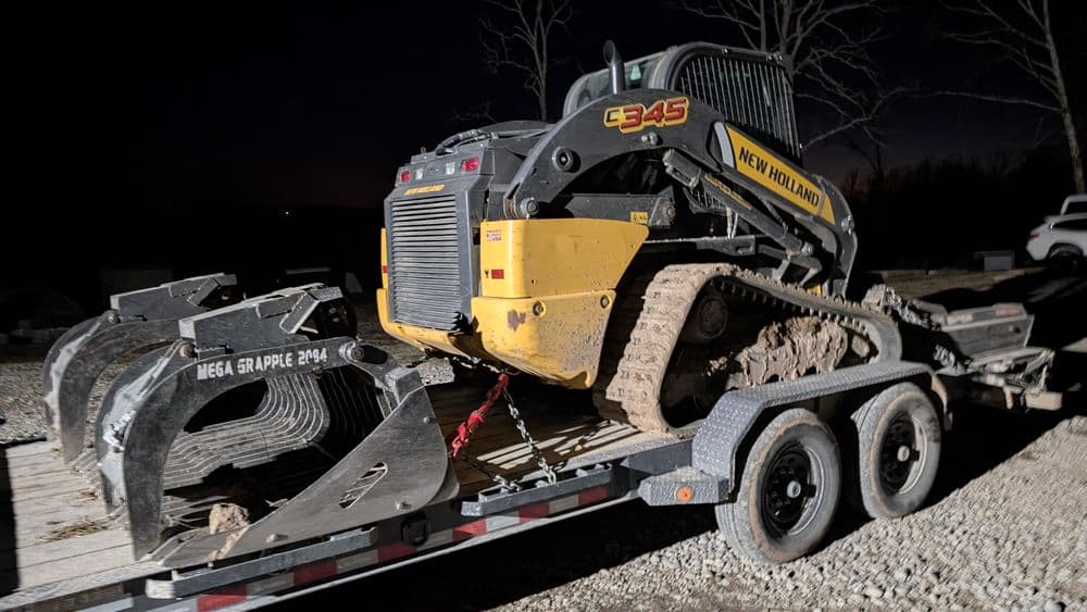 New Holland C345 skid steer with mega grapple attachment on a trailer at night.