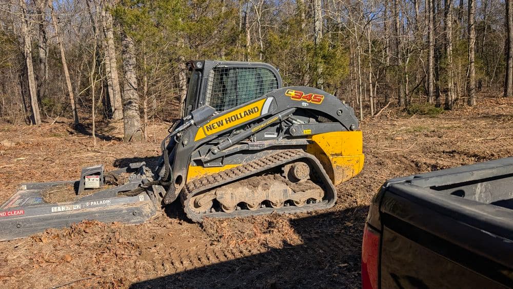 New Holland 345 compact track loader on forested construction site with dirt and trees.