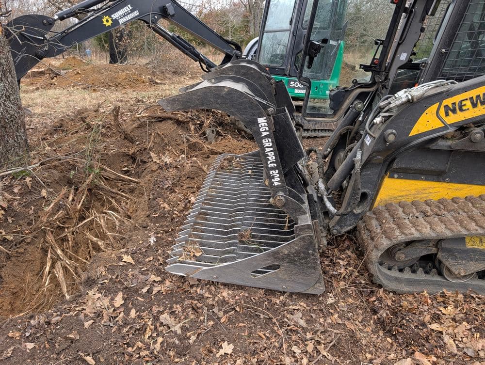 Excavator equipped with a grapple bucket digging near tree roots in a wooded area.