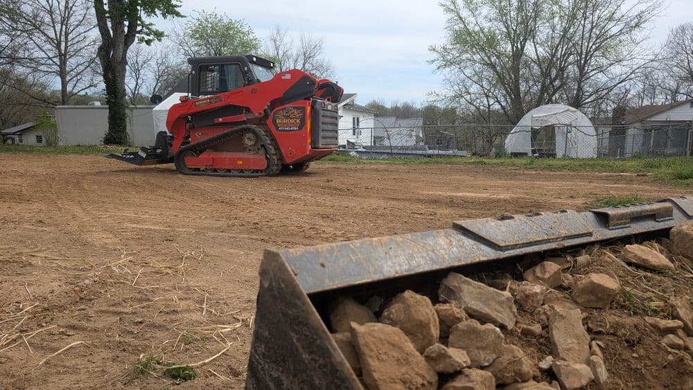 Red compact track loader leveling dirt at a construction site with rocks in the foreground.