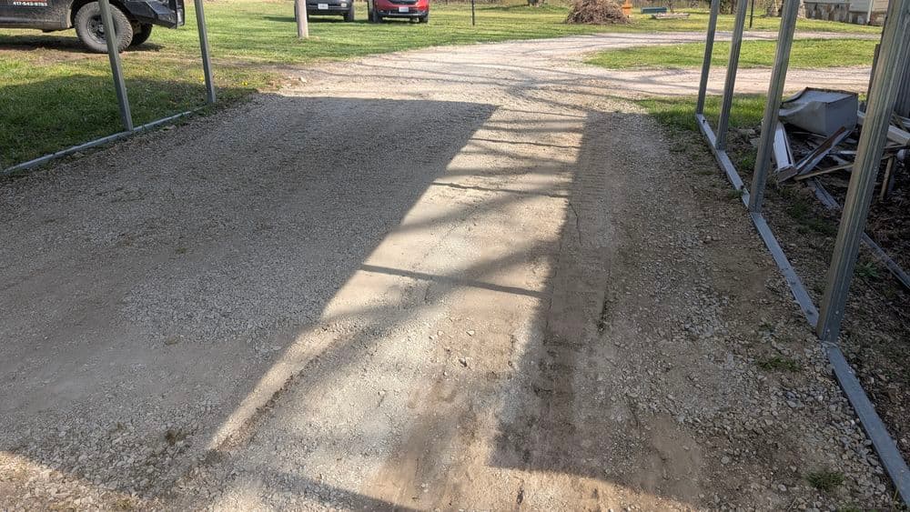 Gravel path with shadows from a nearby structure, leading to vehicles in a grassy area.