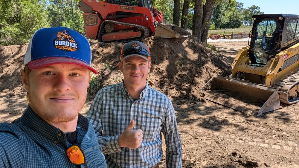 Two men on a construction site with heavy machinery, smiling and giving a thumbs up.
